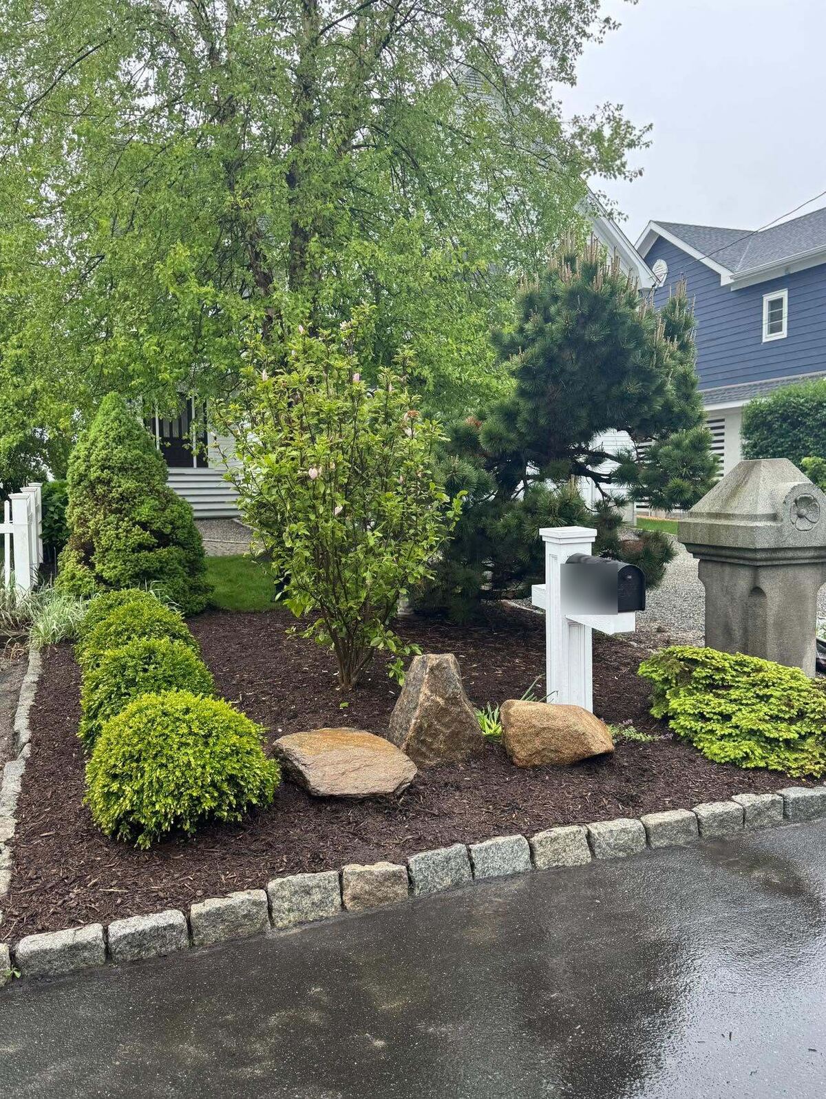 Stone-edged front-yard planting bed with trimmed shrubs, flowers, boulders and a shaped evergreen