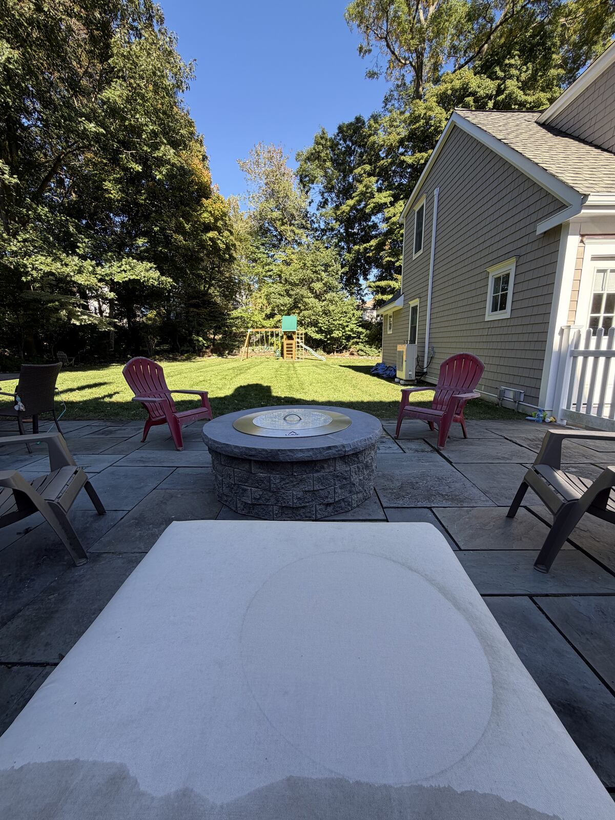 Stone patio with a round fire feature, seating, lawn, and playset beneath leafy trees